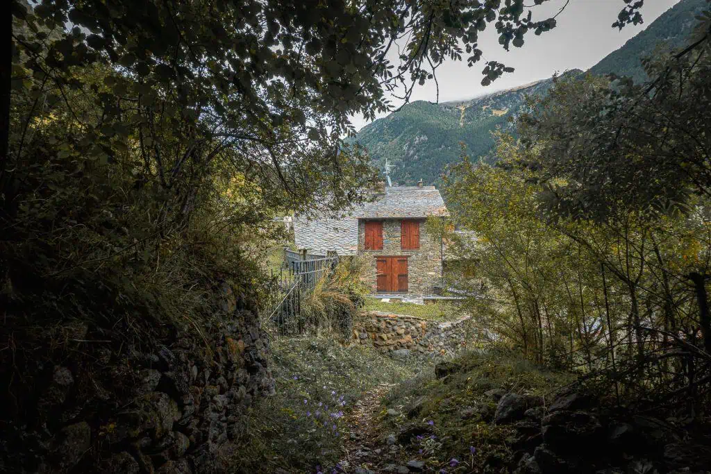 A stone house with red shutters sits nestled among green trees and bushes at the foot of a forested mountain, viewed from a rocky, narrow path along the Pyrenees High Route (HRP), surrounded by lush foliage—perfect for any photo gallery.