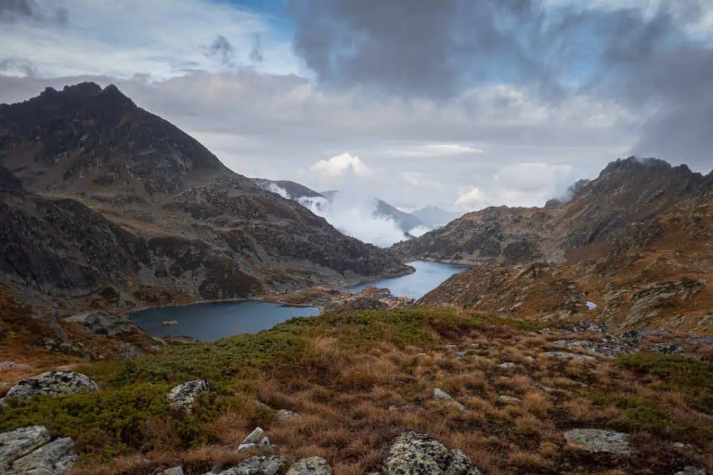 Mountain landscape along the Pyrenees High Route (HRP) featuring rugged peaks, a winding lake, rocky terrain, and patches of grass beneath a cloudy sky—perfect for any photo gallery.