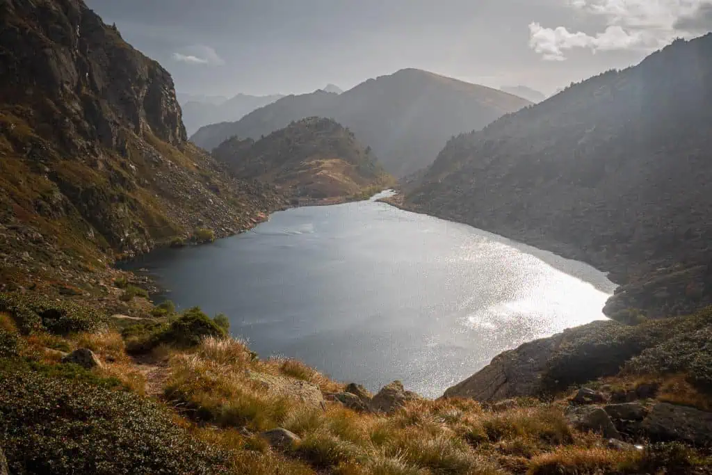 A serene mountain lake reflects sunlight, surrounded by rocky hills and grassy slopes along the Pyrenees High Route (HRP), with distant mountains visible under a partly cloudy sky—an ideal addition to any photo gallery.