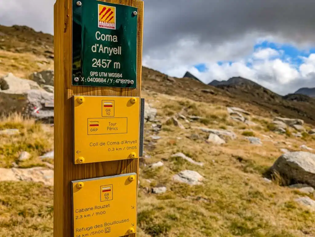 A wooden trail signpost in a mountainous landscape marks hiking routes along the Pyrenees High Route (HRP) at Coma d’Anyell, 2457 meters high, under a partly cloudy sky. Rocky hills and grassy terrain stretch out in the background.