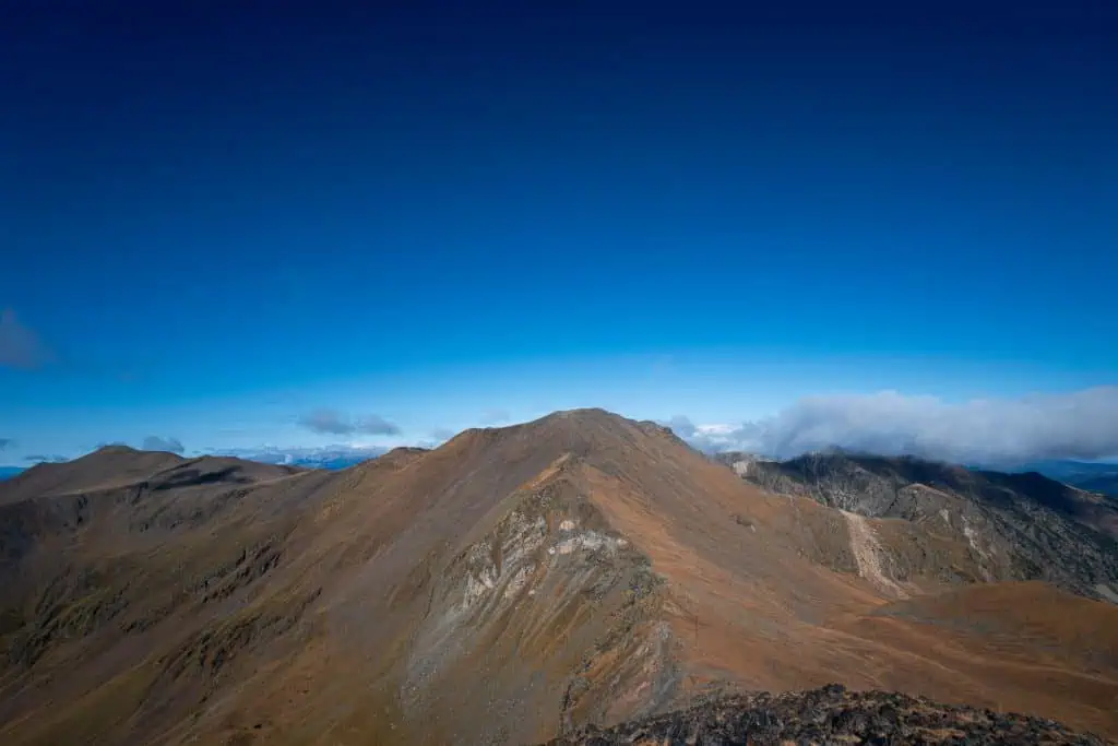 Wide-angle view of brown, rocky mountain ridges under a clear blue sky with scattered clouds in the distance. Sunlight casts shadows across the rugged landscape along the Pyrenees High Route (HRP) for a stunning photo gallery scene.