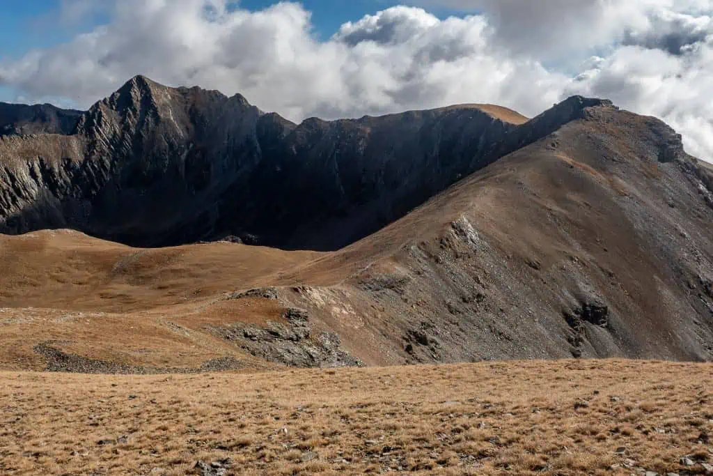 Mountain range under a partly cloudy sky, with rugged, rocky peaks and slopes covered in dry, golden grass in the foreground—an iconic view along the HRP (Pyrenees High Route), perfect for any photo gallery. Light reveals hill contours and deep mountain shadows.