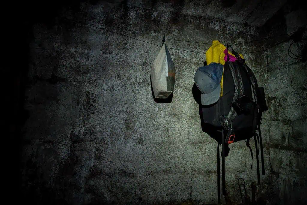 A dimly lit, rough concrete wall displays a plastic bag, backpack, jacket, and cap hanging from hooks—evoking the worn, moody atmosphere of a Pyrenees High Route (HRP) rest stop. Perfect for a Photo Gallery with stories of rugged adventure.
