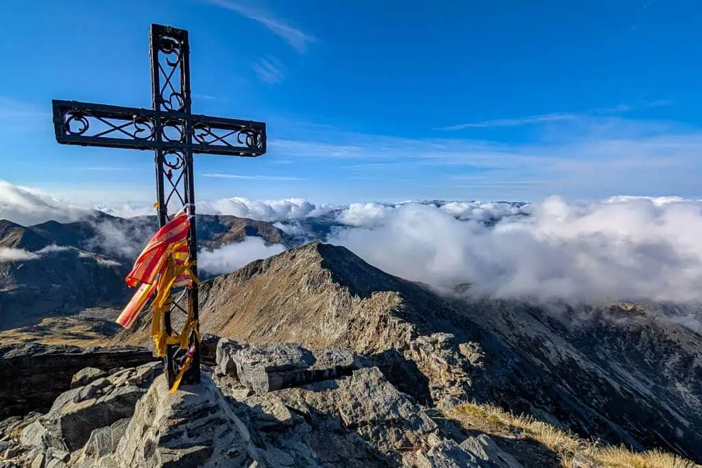 A metal cross with colorful ribbons stands on a rocky mountain peak along the Pyrenees High Route, overlooking distant ridges and valleys beneath a bright blue sky with scattered clouds.