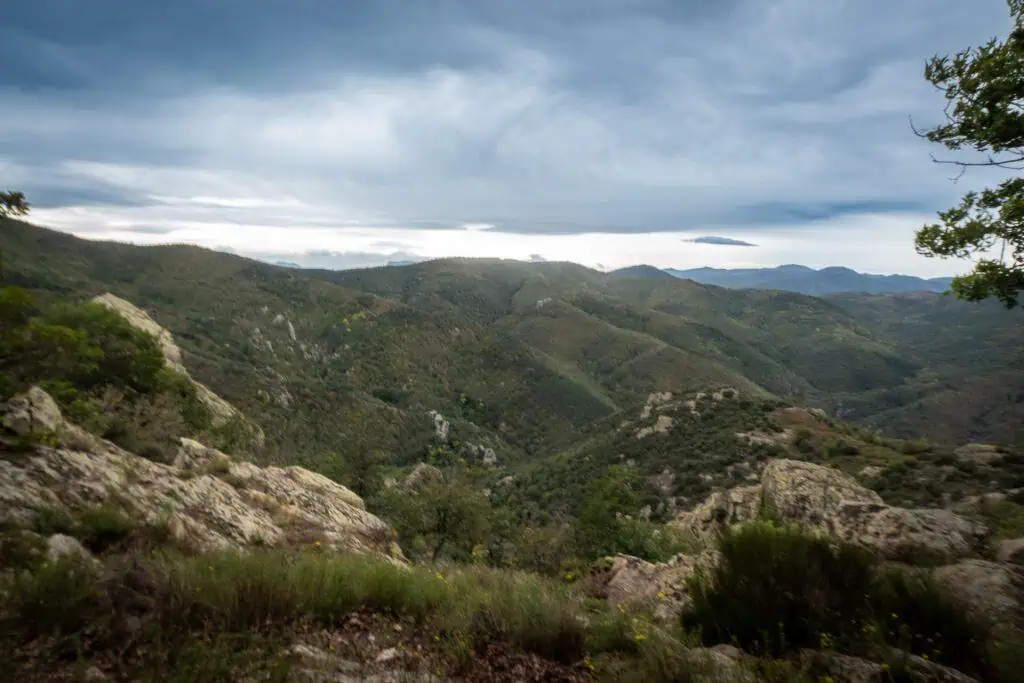 A wide view of rolling green hills and rocky terrain under a cloudy, overcast sky, with mountains visible in the distance along the Pyrenees High Route (HRP).