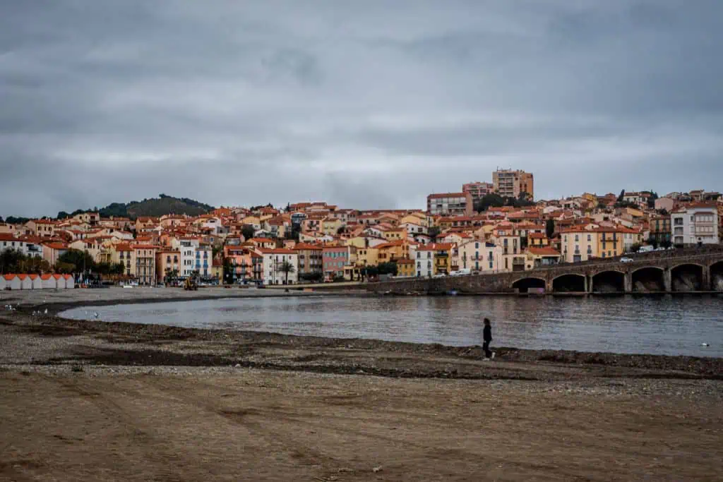 A person walks alone on a deserted beach under cloudy skies, with colorful buildings and a stone bridge in the background, capturing a scene worthy of any photo gallery along the shoreline of a coastal town.