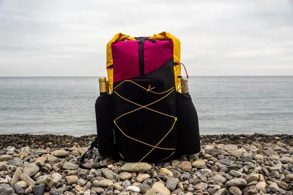 A colorful backpack with yellow and magenta accents sits on a rocky beach along the Pyrenees High Route, with the sea and cloudy sky in the background. Two water bottles are visible in side pockets—perfect for your HRP photo gallery.