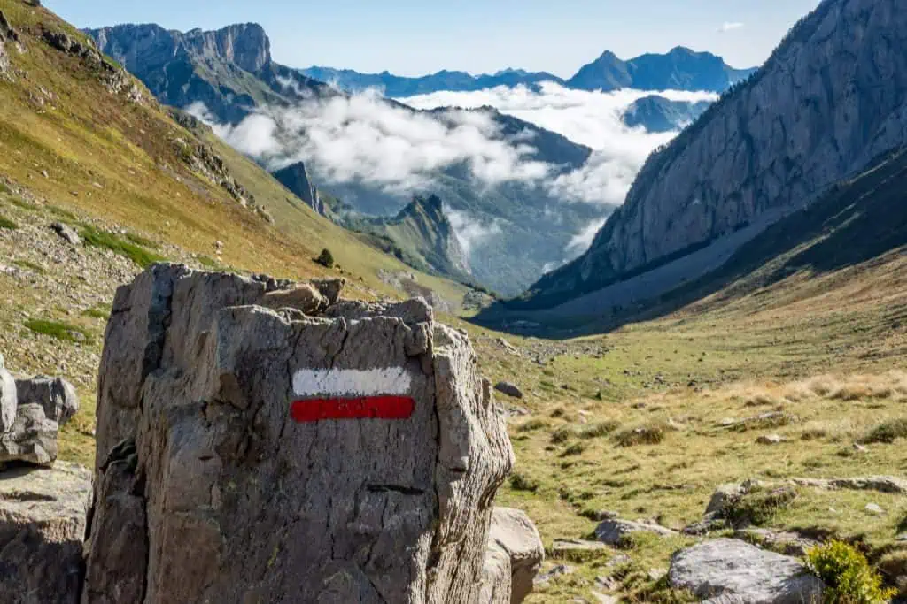 A large rock marked with red and white stripes sits in the foreground of a scenic mountain valley, with rugged peaks, green slopes, and clouds settled in the distant landscape.