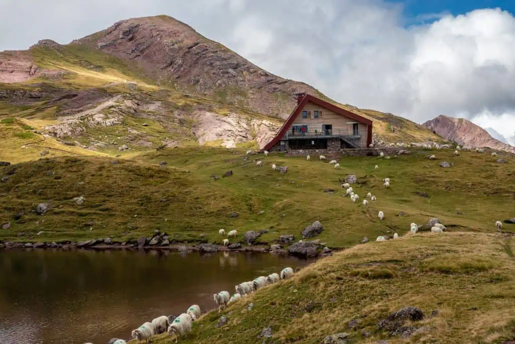 A wooden cabin sits on a grassy hill near a pond, surrounded by grazing sheep. Rocky mountains rise in the background under partly cloudy skies.