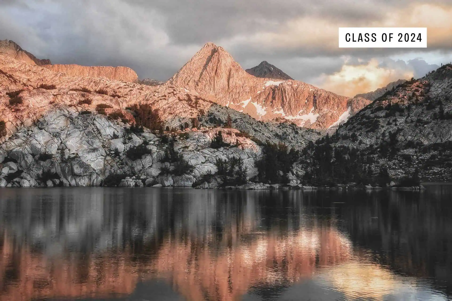 Rocky mountains with snow patches reflected in a calm lake at sunset, illuminated by warm golden light. A label in the sky reads Class of 2024, inspired by epic JMT horror stories 2024 along the trail.