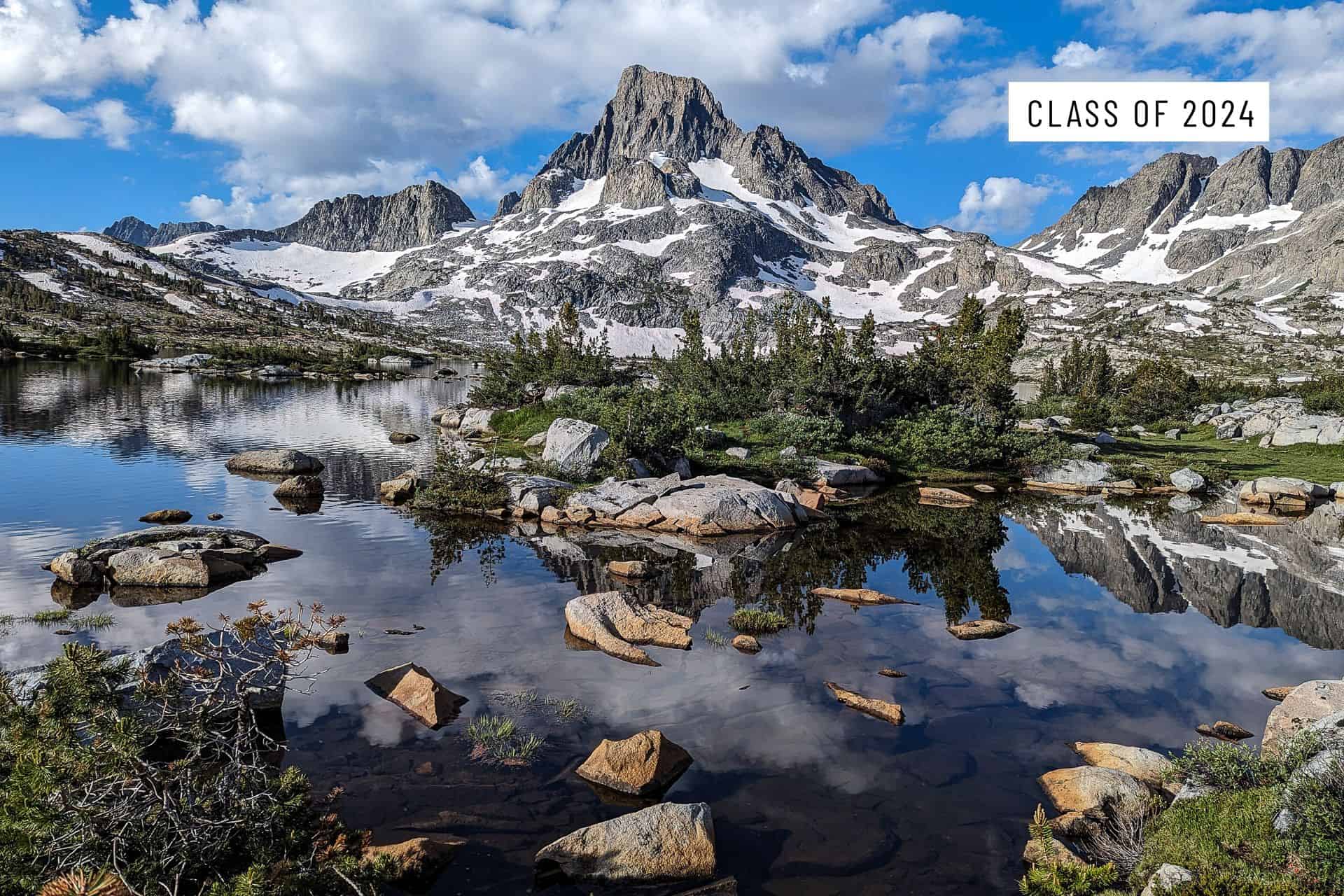 Snow-capped mountains reflected in a calm alpine lake with scattered rocks and trees. In the sky, text reads Class of 2024. Puffy clouds float above the rugged peaks—memories of JMT worst moments 2024 fade under a bright blue sky.