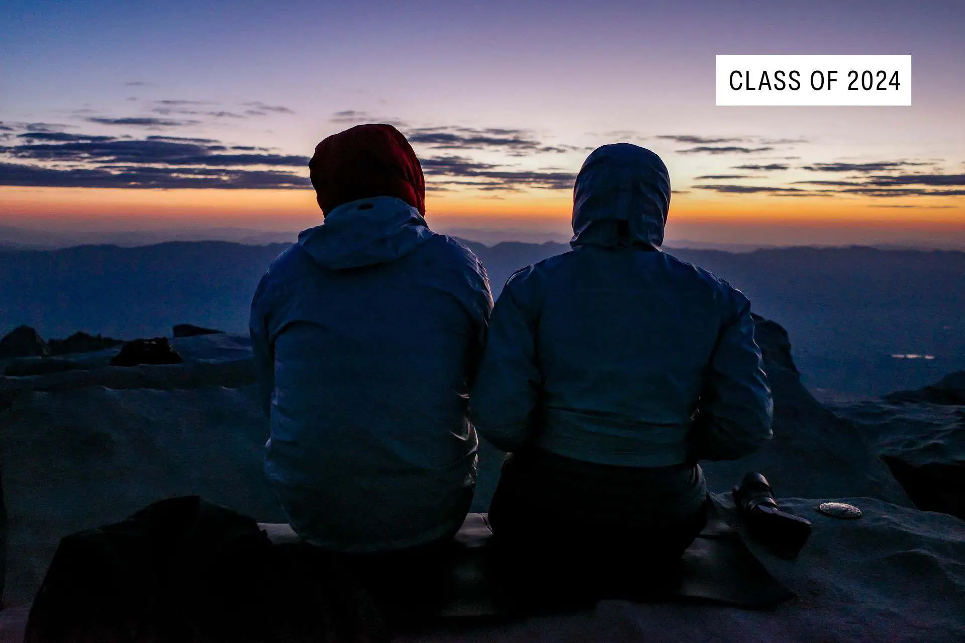 Two people in hooded jackets sit on a mountain top at sunrise, gazing at the horizon. A sign in the sky reads Class of 2024—this peaceful scene is a stark contrast to any JMT horror stories 2024 may bring. Cool blue and orange tones fill the sky.