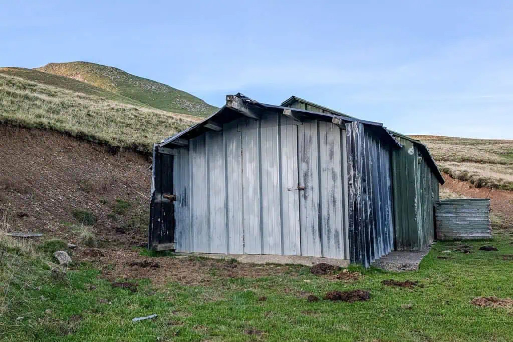 A weathered metal shed, reminiscent of HRP shelters part 1, stands on grassy terrain with hills in the background under a clear blue sky. The shed appears old and worn, its paint faded and peeling.