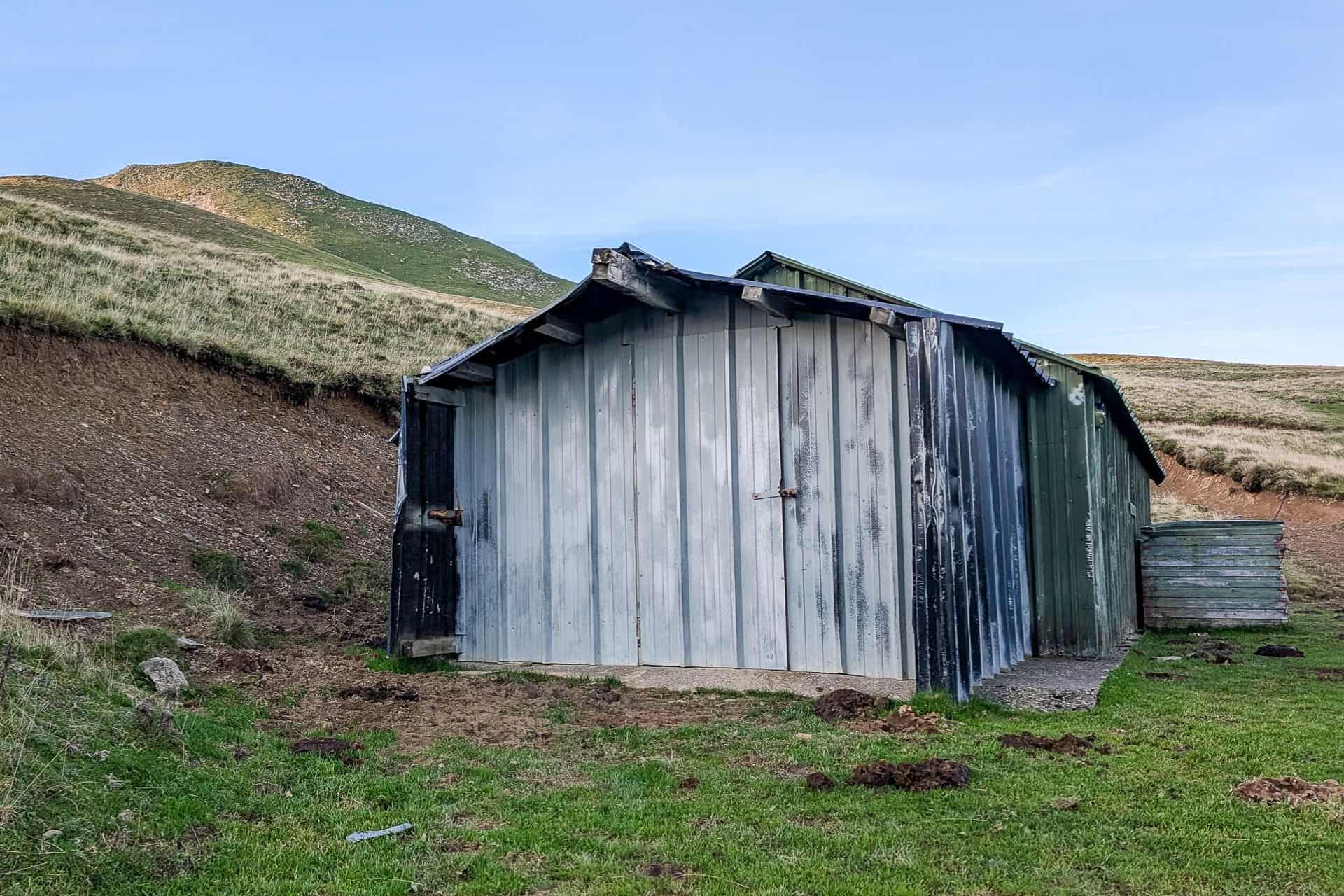 A weathered metal shed, reminiscent of HRP shelters part 1, stands on grassy terrain with hills in the background under a clear blue sky. The shed appears old and worn, its paint faded and peeling.