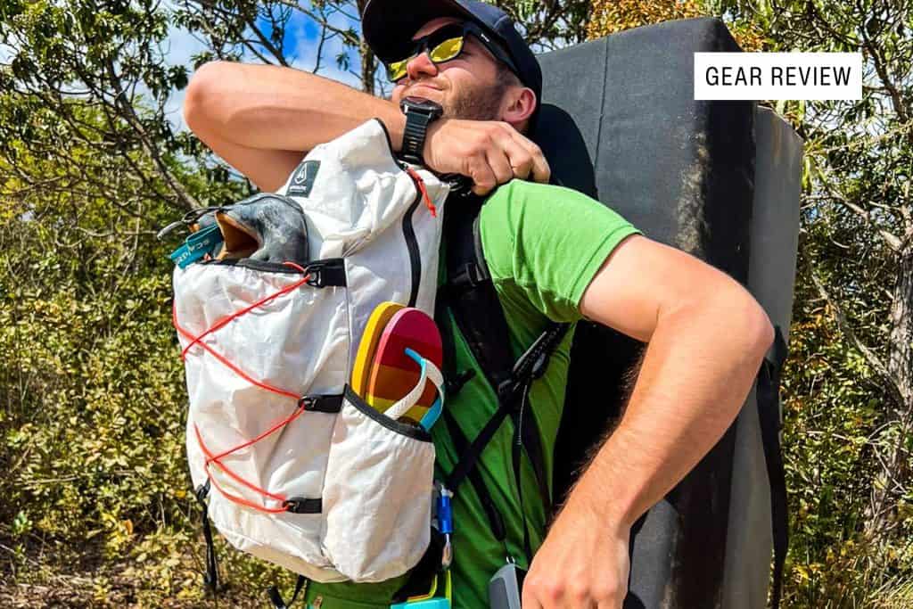 A smiling man outdoors adjusts his white Hyperlite Mountain Gear Daybreak backpack, stuffed with climbing gear. He wears sunglasses, a green shirt, and a cap, with a crash pad on his back. Trees and bushes surround him. A “Gear Review” label is in the corner.