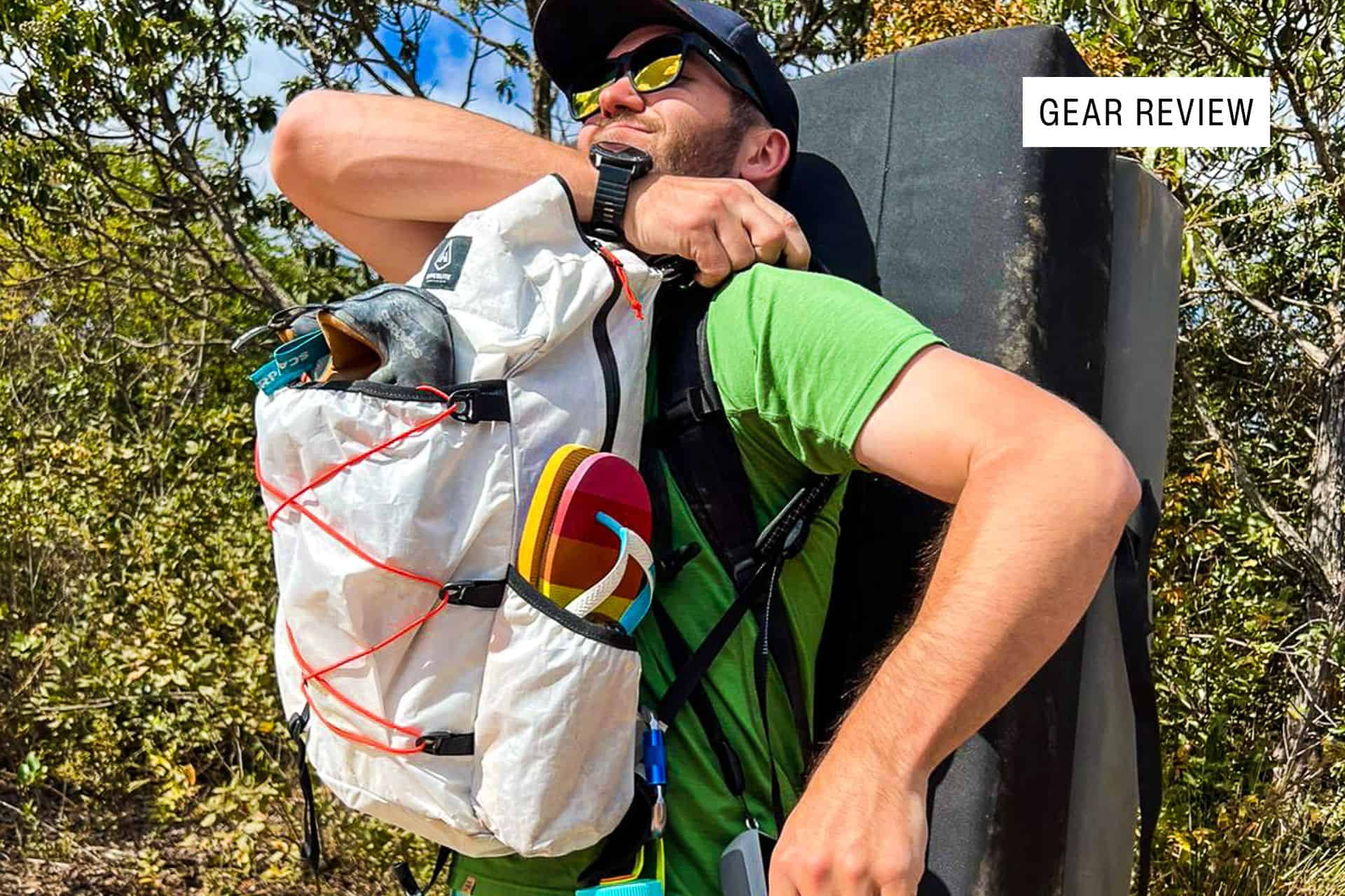 A smiling man outdoors adjusts his white Hyperlite Mountain Gear Daybreak backpack, stuffed with climbing gear. He wears sunglasses, a green shirt, and a cap, with a crash pad on his back. Trees and bushes surround him. A “Gear Review” label is in the corner.