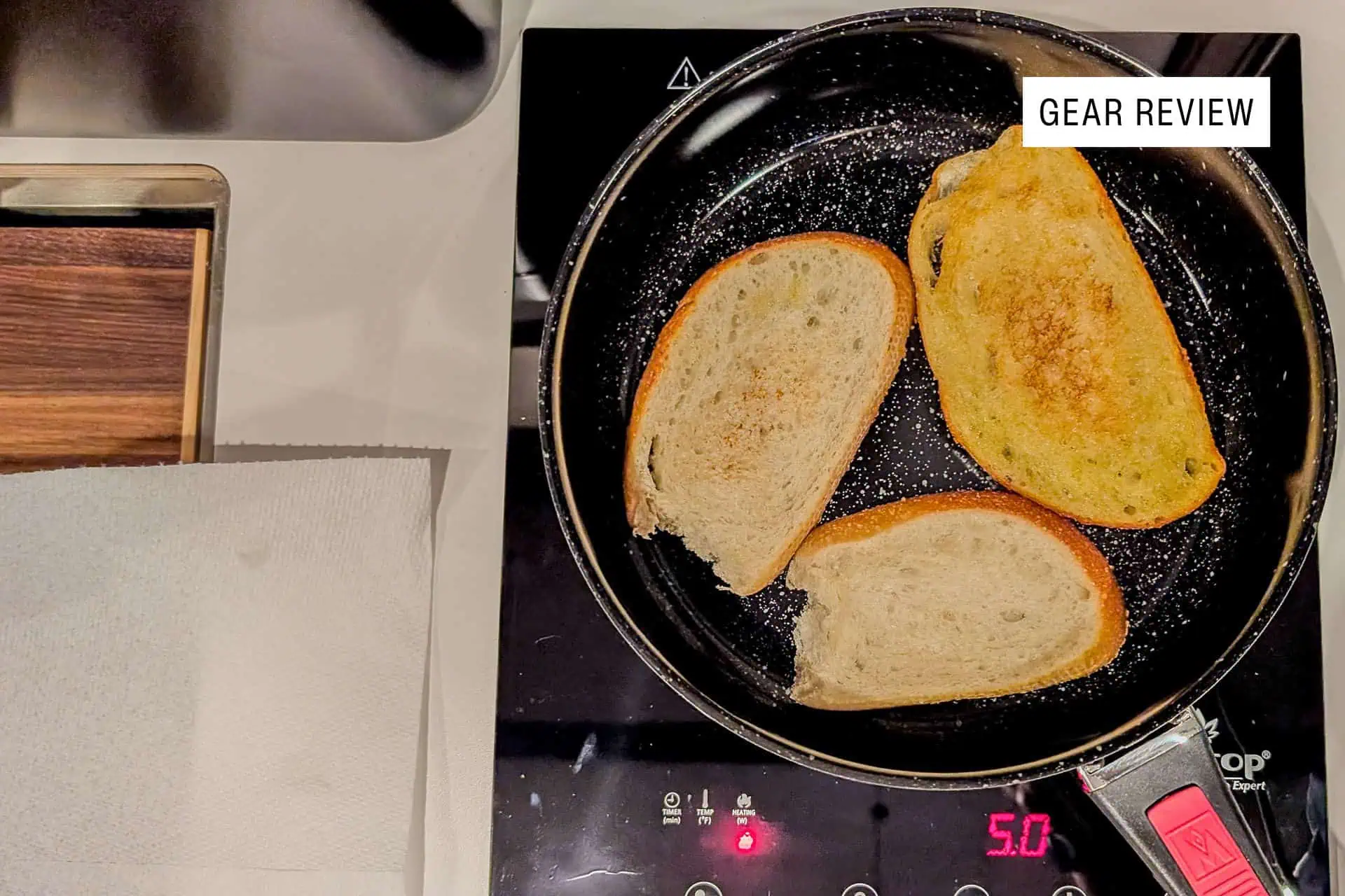 Three slices of bread are toasting in a frying pan on an induction stove set to medium heat. A paper towel is on the counter nearby, and a label in the corner reads Gear Review: Hyperlite Mountain Gear Daybreak.