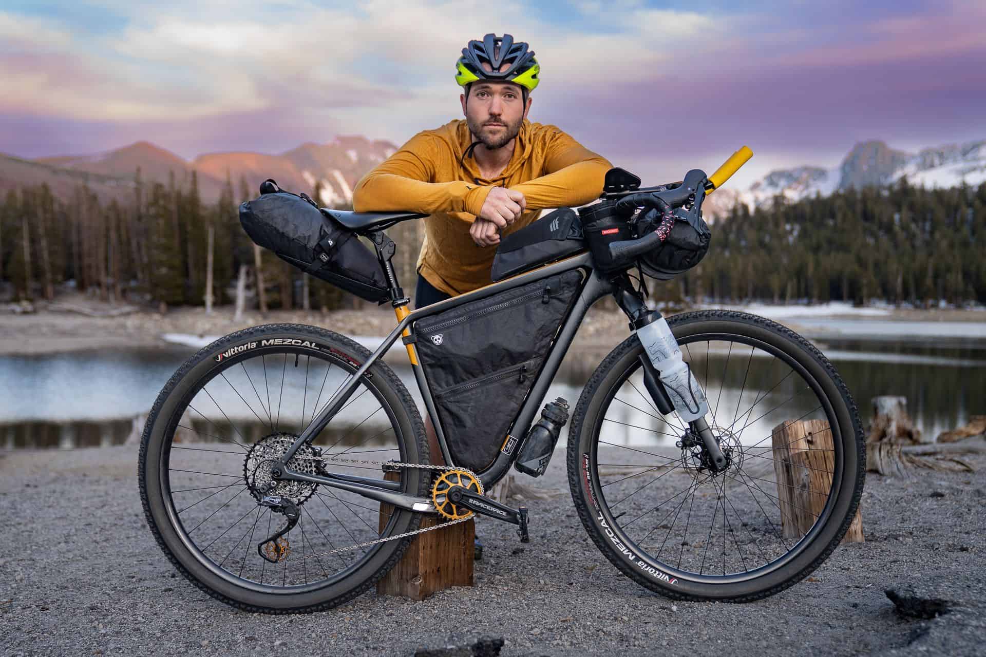 A man in a yellow jacket and helmet leans on his loaded touring bike, fresh from the Tour Divide, parked by a lake with mountains and pine trees in the sunset—perfect for a post race report.