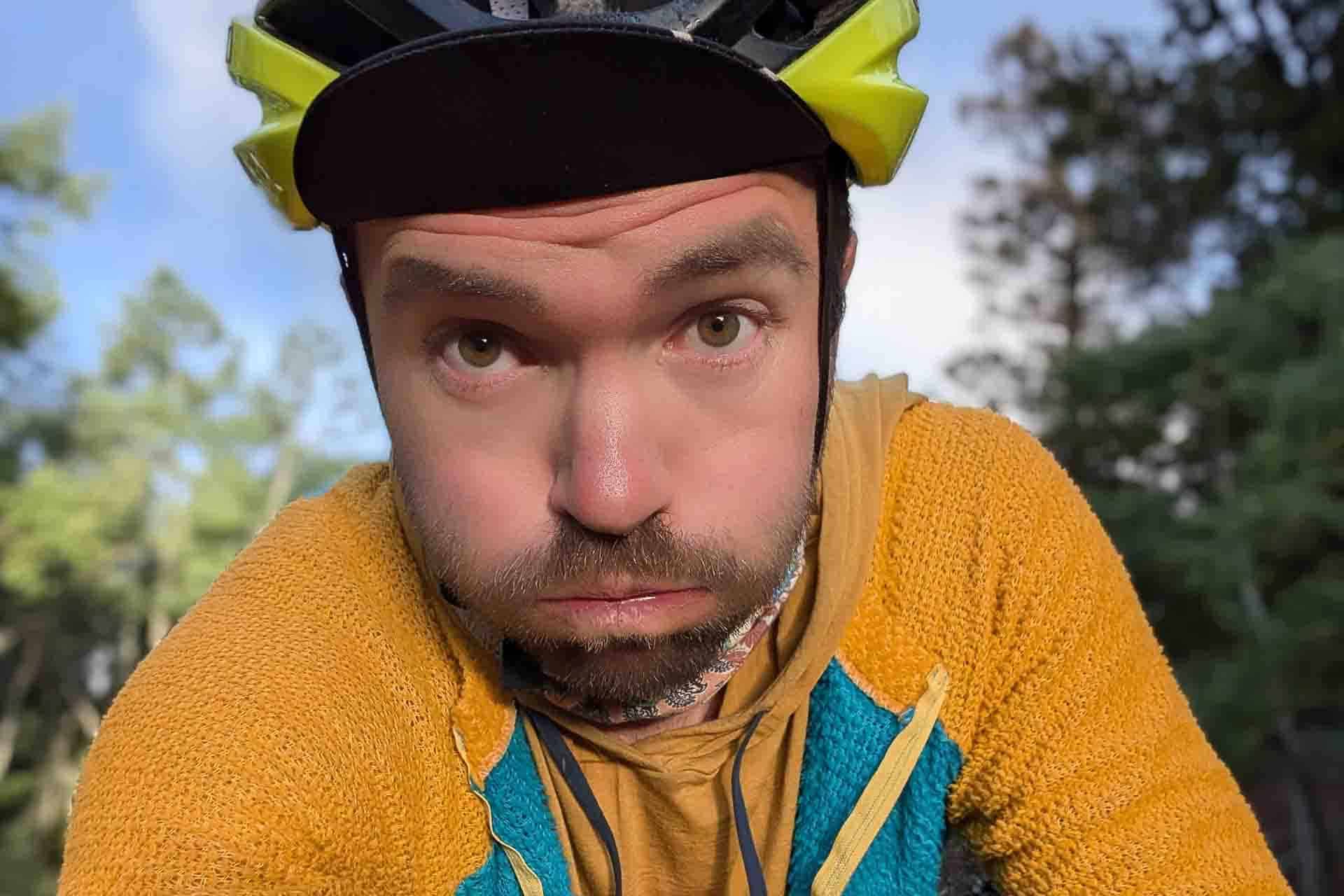 A man wearing a bike helmet and a yellow-green jacket looks directly at the camera with puffed cheeks, perhaps reflecting on Tour Divide changes. Trees and blue sky are blurred in the background.