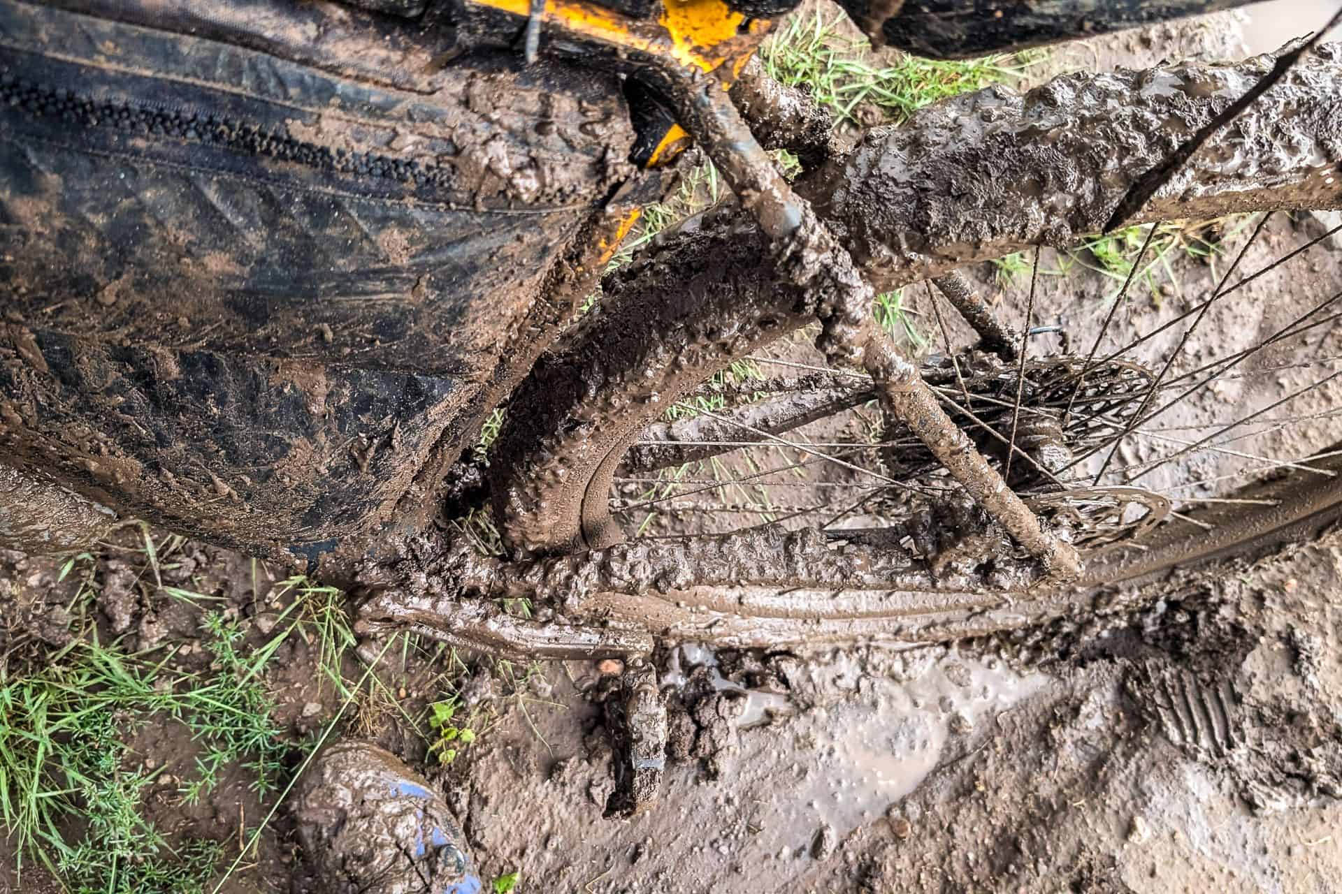 A bicycle wheel and frame, caked in thick, wet mud from the tour divide, with grass and muddy ground nearby—mud obscuring most bike details and hinting at what went wrong on this rugged journey.