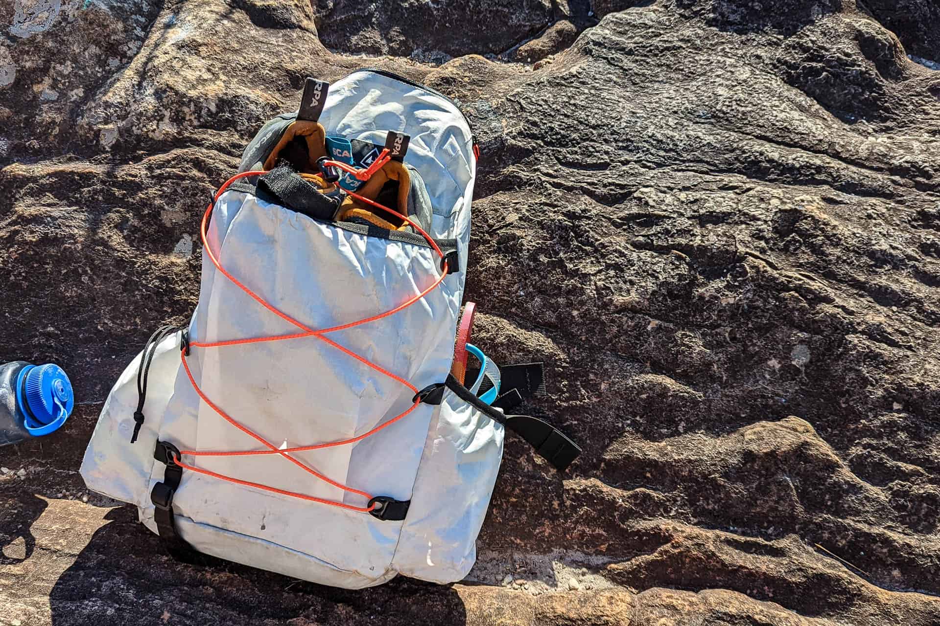 A white Hyperlite Mountain Gear Daybreak backpack with orange bungee cords and climbing gear sits on a rocky surface in sunlight. Part of a blue water bottle is visible on the left edge of the image.