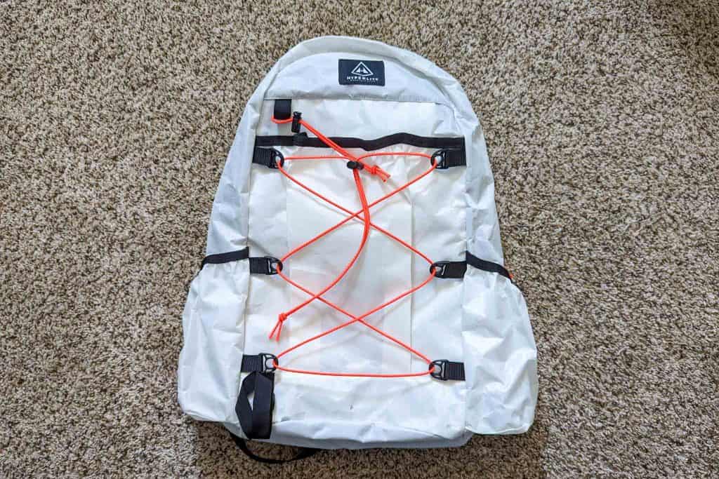 A white hiking backpack with black buckles and bright orange bungee cords lies flat on a light brown carpeted floor beside a Rogue Panda Blue Ridge Handlebar Harness.