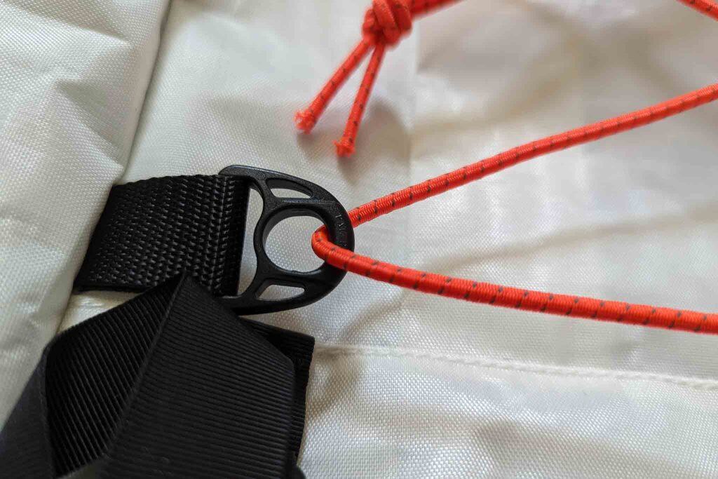 Close-up of an orange elastic cord threaded through a black plastic buckle, attached to black webbing on a white fabric surface—part of the Rogue Panda Blue Ridge Handlebar Harness. Another orange cord is visible in the background.