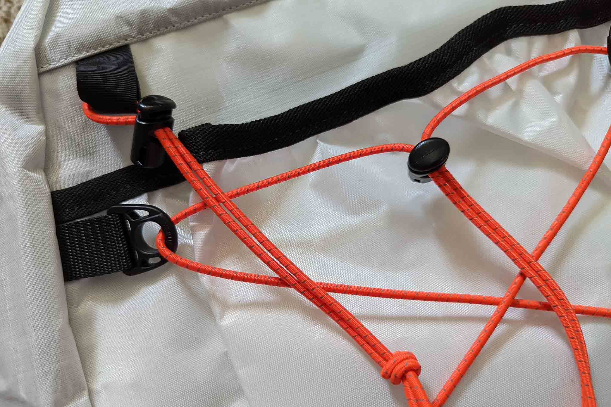 Close-up of orange elastic bungee cords with black plastic clips and sliders attached to a light gray fabric surface, likely part of a Rogue Panda Blue Ridge Handlebar Harness or similar outdoor gear.