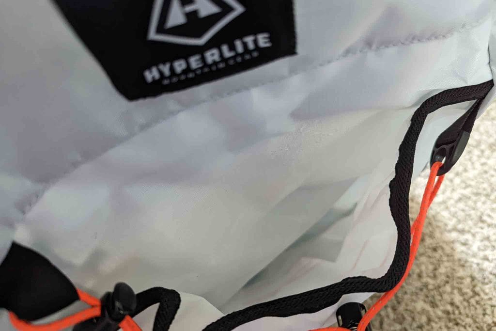 Close-up of a light gray Hyperlite backpack showing part of the logo, black trim, and an orange drawstring beside a Rogue Panda Blue Ridge Handlebar Harness on a carpeted floor.