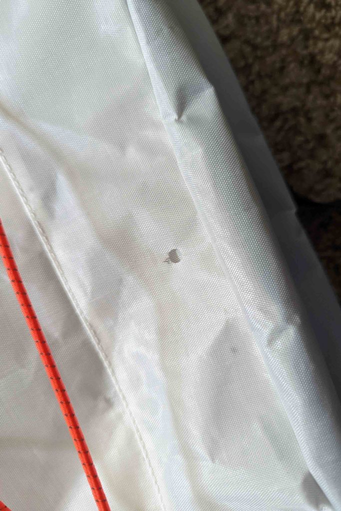 A close-up of white fabric with a small hole in the center, featuring red stitching running vertically along the left—part of a Rogue Panda Blue Ridge Handlebar Harness against a darker, textured background.