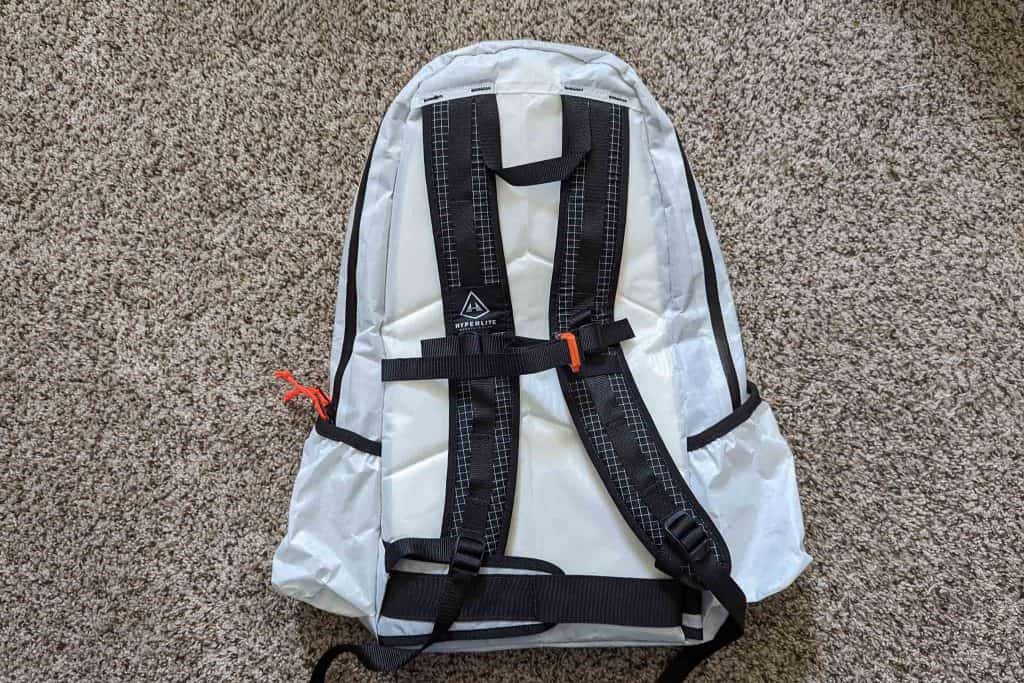 A white backpack with black straps and trim lies on a beige carpet. It features a small orange clip on one strap, a visible logo on the back panel, and resembles the sleek design of the Rogue Panda Blue Ridge Handlebar Harness.