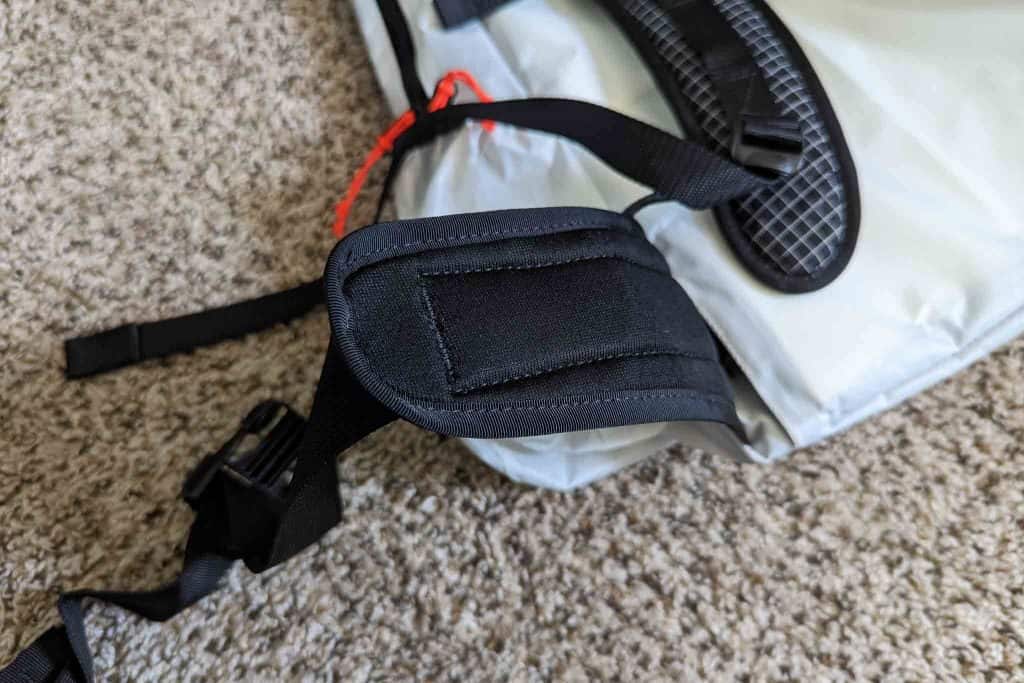 A close-up of a black padded strap attached to a gray bag, likely part of a Rogue Panda Blue Ridge Handlebar Harness, lying on a beige carpet. A red cord is visible near the top of the image.