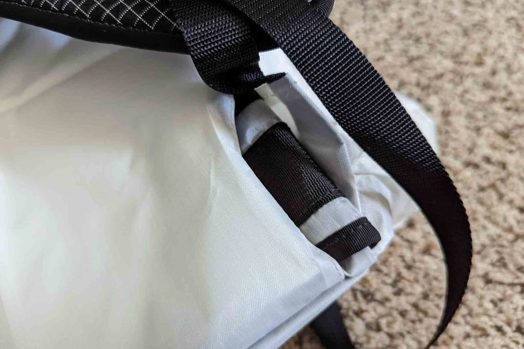 Close-up of a white and black backpack with black straps, showing a partially inserted black item in a side pocket—resting on light brown carpet next to a Rogue Panda Blue Ridge Handlebar Harness.