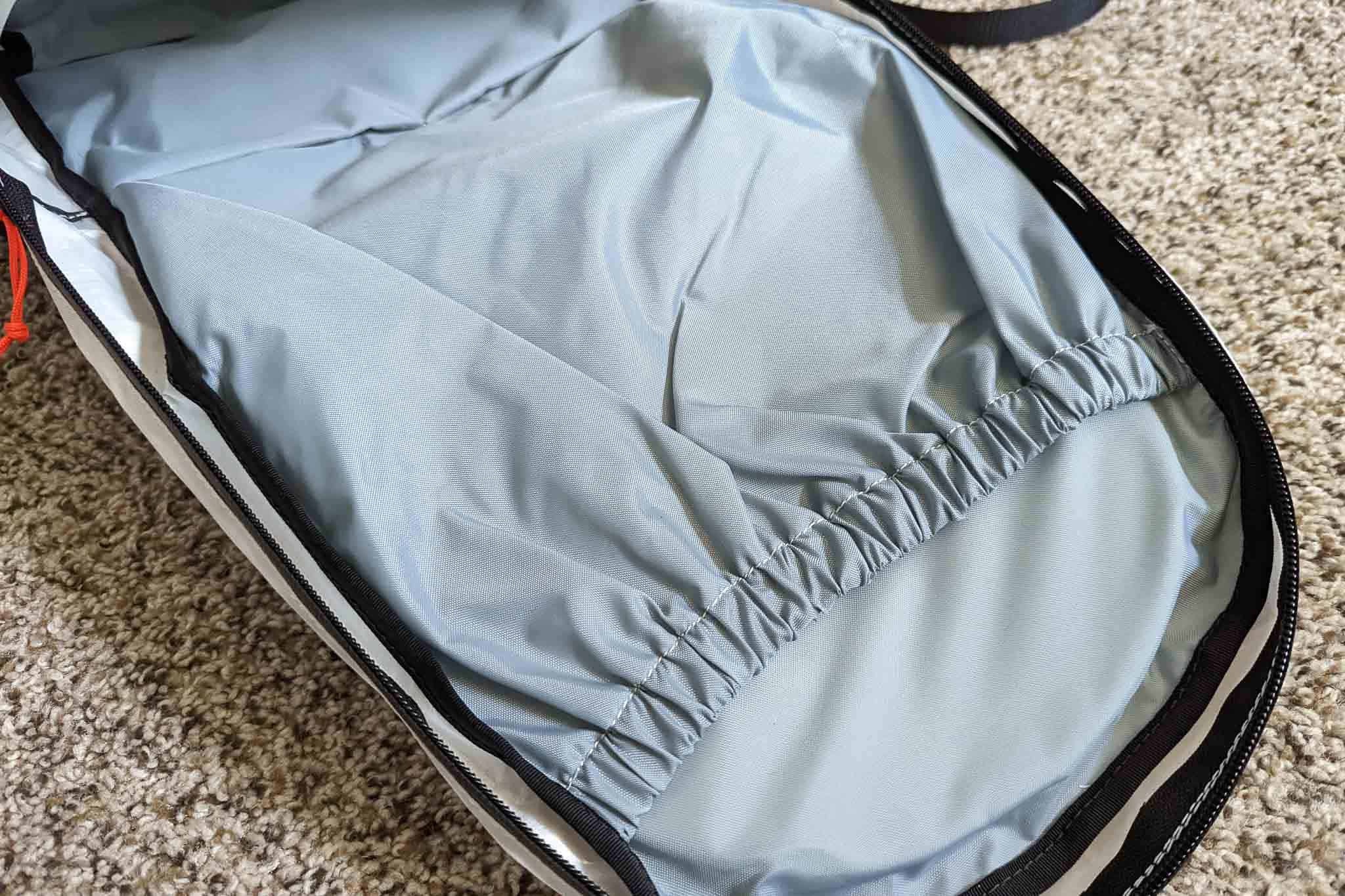 A close-up of the inside of an open backpack, paired with a Rogue Panda Blue Ridge Handlebar Harness, shows a light gray, smooth fabric lining with an internal elastic pocket, resting on a beige carpeted floor.
