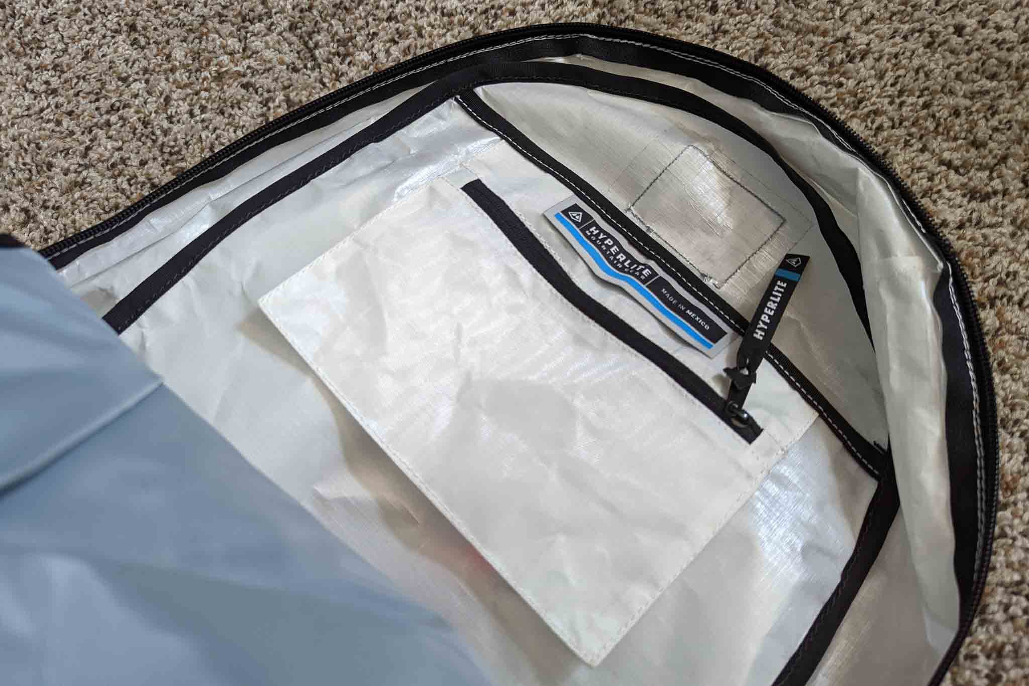 A close-up of the inside of a white travel bag, showing a zippered mesh pocket labeled Peak Design lying on a beige carpet. The pocket features a black zipper, a blue-accented branded tag, and space to fit gear like the Rogue Panda Blue Ridge Handlebar Harness.