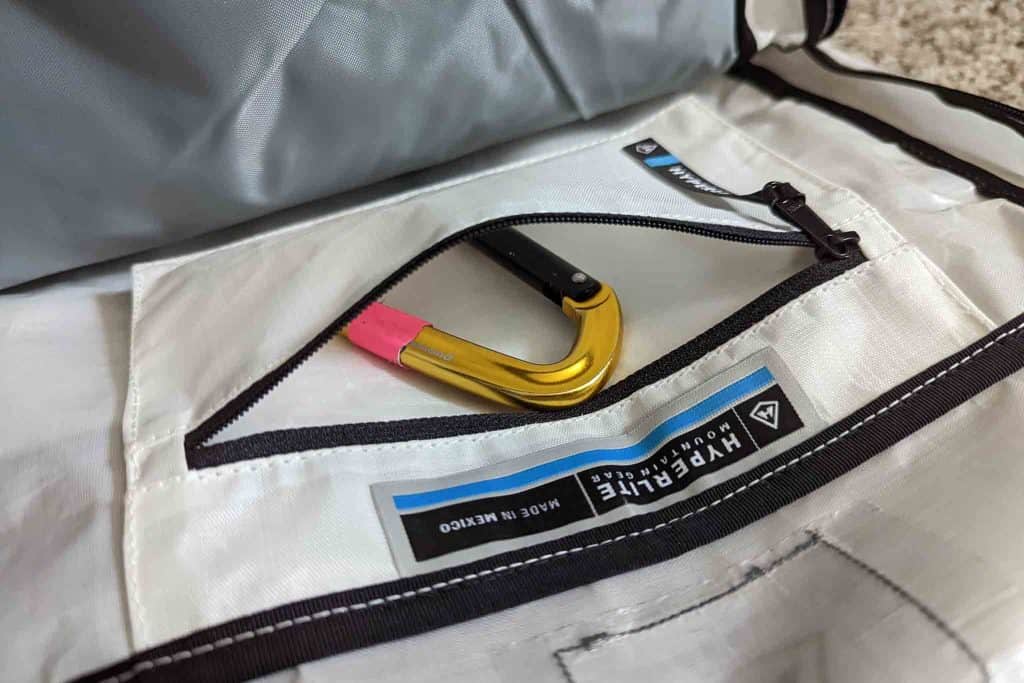 A gold carabiner with a pink section rests inside a white zippered pocket of a bag labeled HYPERLITE, featuring black trim and stitching. The setup pairs well with the Rogue Panda Blue Ridge Handlebar Harness for organized adventures.