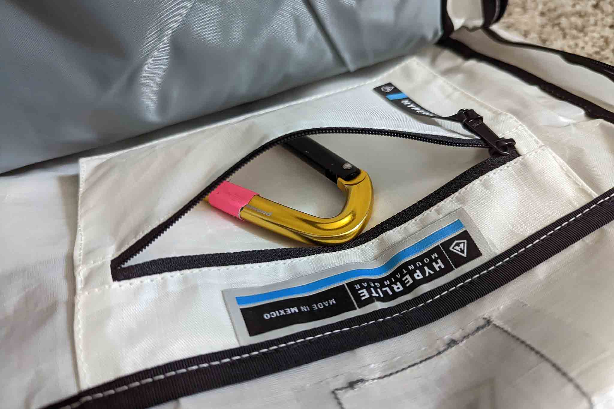 A gold carabiner with a pink section rests inside a white zippered pocket of a bag labeled HYPERLITE, featuring black trim and stitching. The setup pairs well with the Rogue Panda Blue Ridge Handlebar Harness for organized adventures.