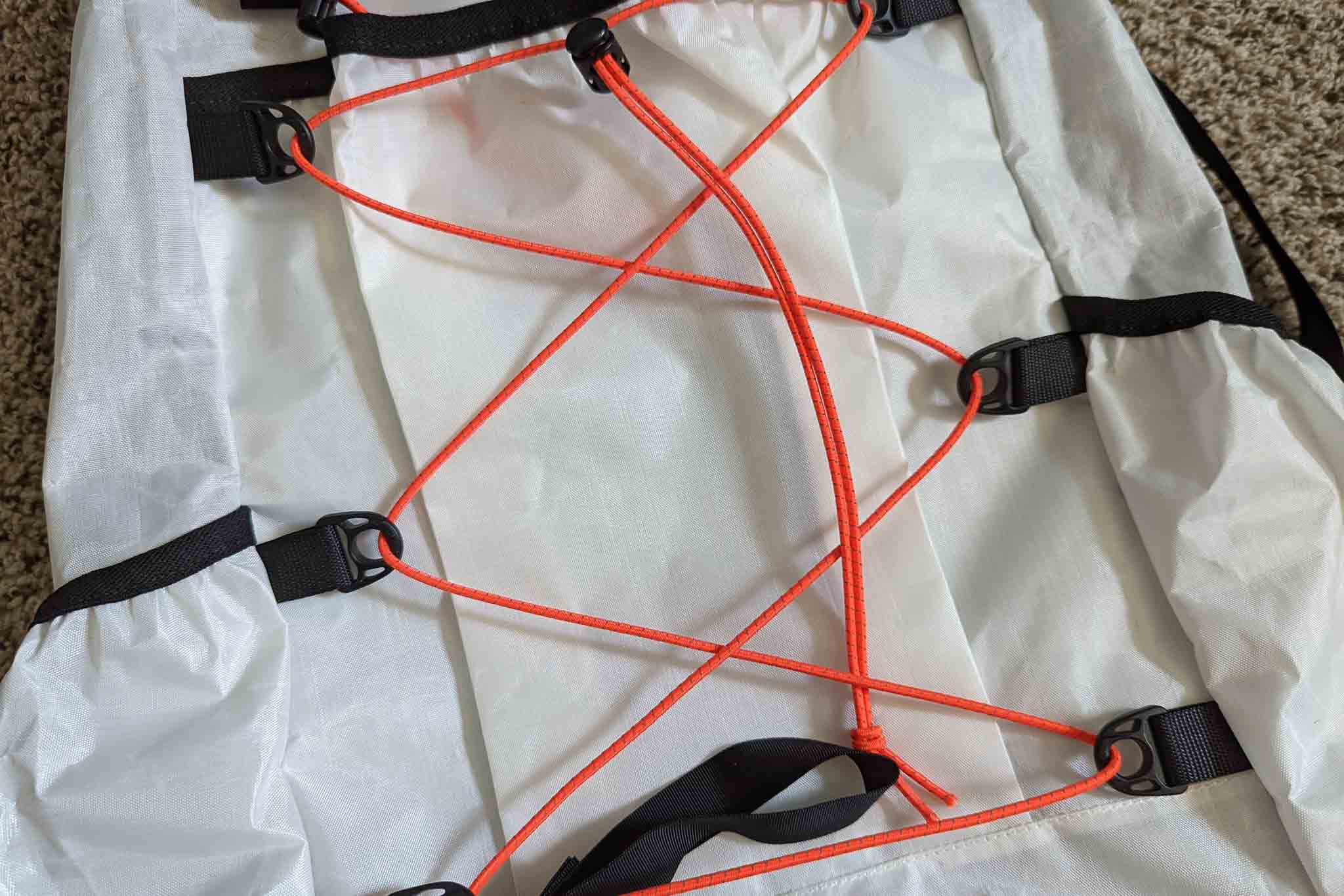 Close-up of orange elastic cords crisscrossed and secured by black loops on a white fabric surface, likely part of a Rogue Panda Blue Ridge Handlebar Harness, lying on a carpeted floor.