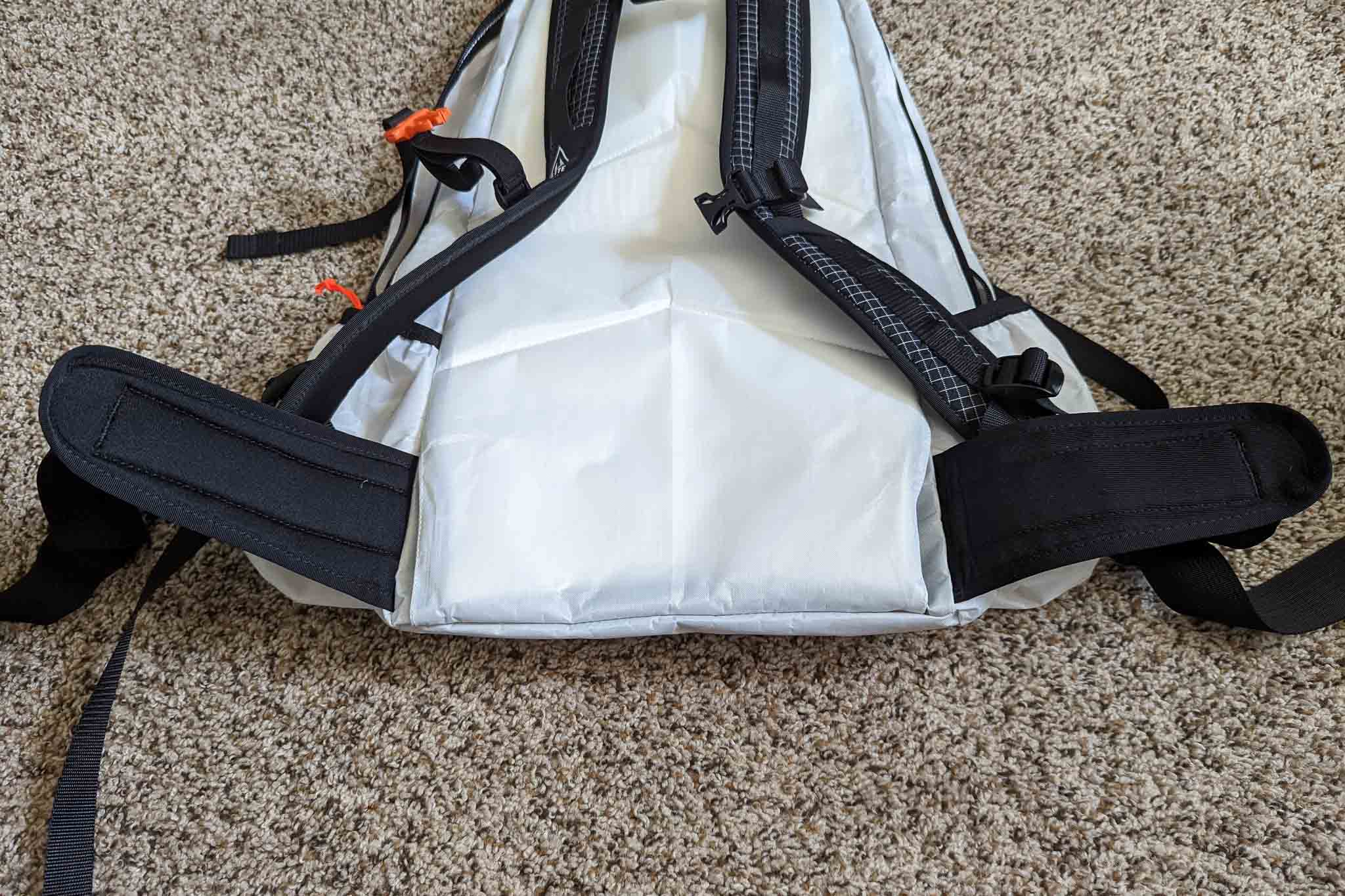 A close-up of the back side of a white backpack with black shoulder straps and hip belts, lying on a beige carpeted floor. Orange cord accents and visible stitching give it a look similar to the Rogue Panda Blue Ridge Handlebar Harness.