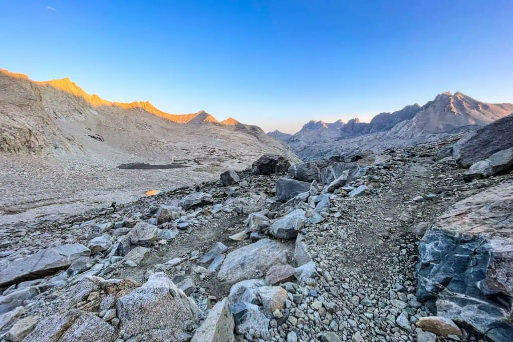 A rocky mountain trail winds through a barren, rugged landscape under a clear blue sky, echoing some of the JMT's most surprising moments of 2024, with distant peaks bathed in early morning sunlight.