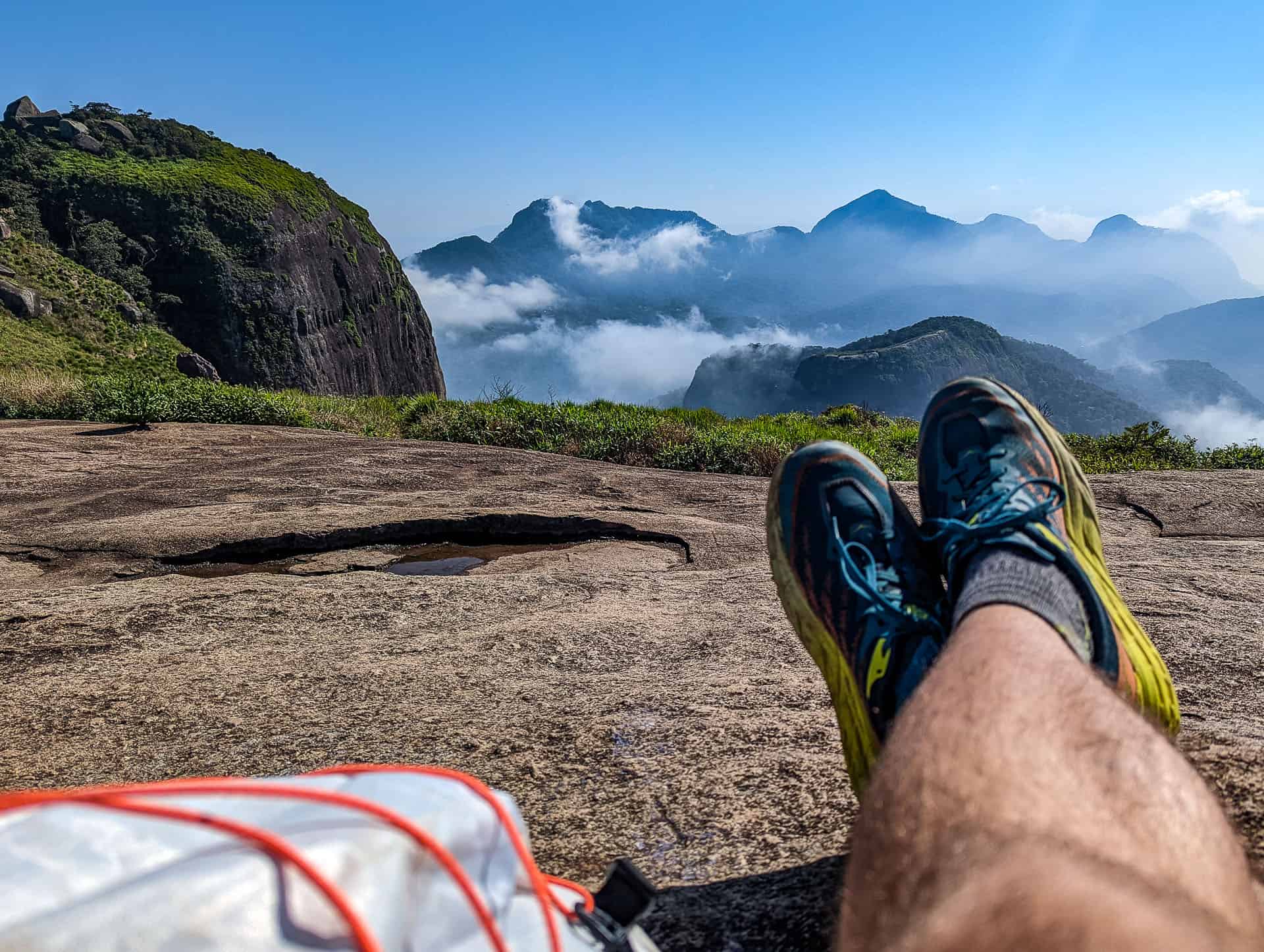 A person’s legs and feet in running shoes rest on a rocky surface, overlooking misty mountains and lush greenery beneath a bright blue sky. A Hyperlite Mountain Gear Daybreak backpack is partially visible in the foreground.