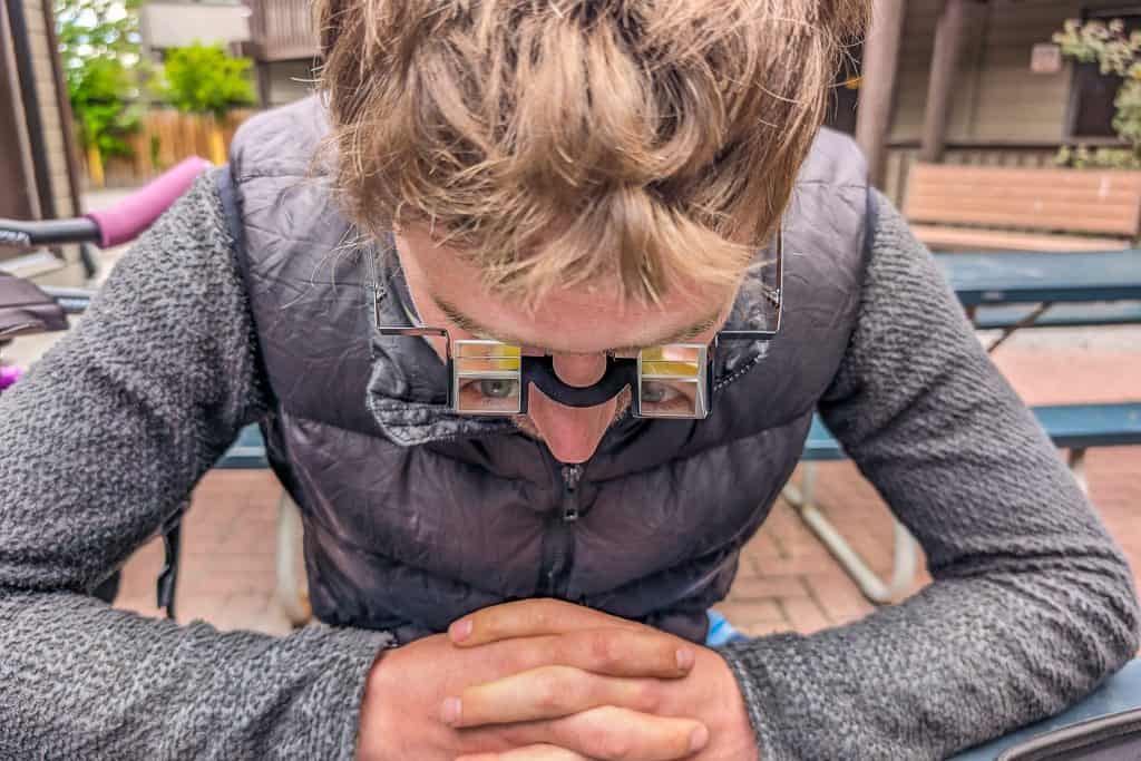 A person with light brown hair, head down and hands clasped, sits at an outdoor table in a gray jacket and mirrored prism glasses—perfect for keeping the tour divide body posture while seeing forward.