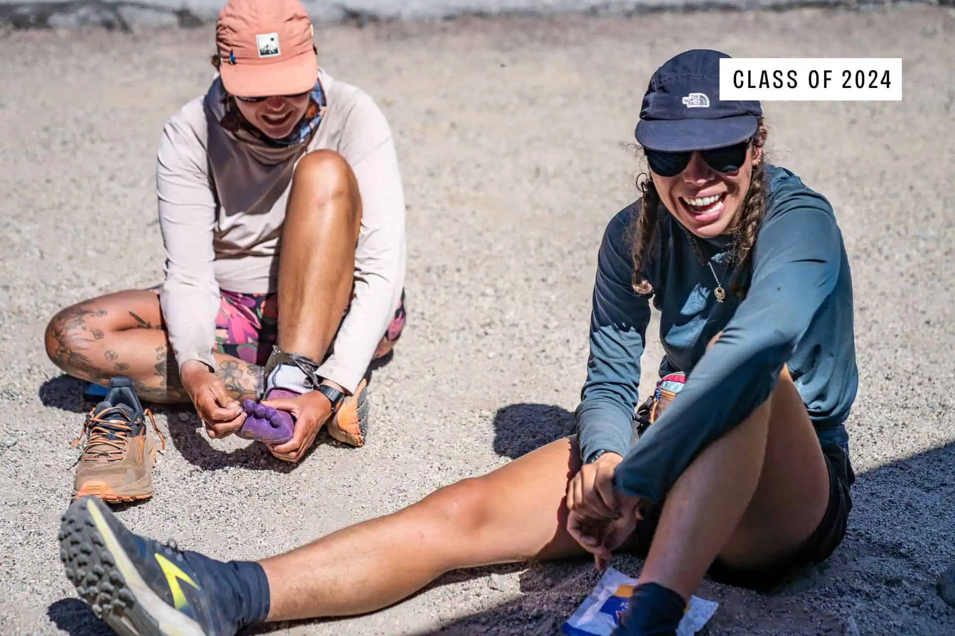 Two hikers sit on the ground smiling and relaxing. One adjusts her sock while the other laughs, both wearing hats and outdoor clothing, showcasing womens PCT gear 2024. The text CLASS OF 2024 appears in the corner.