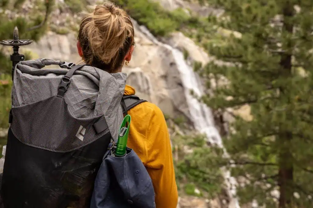 A person with a backpack stands outdoors facing a blurred waterfall and forest. The hiker, perfect for any backpackers Fourth of July sales guide, has blonde hair tied up, wears a yellow jacket, and carries trekking poles and a green water bottle.