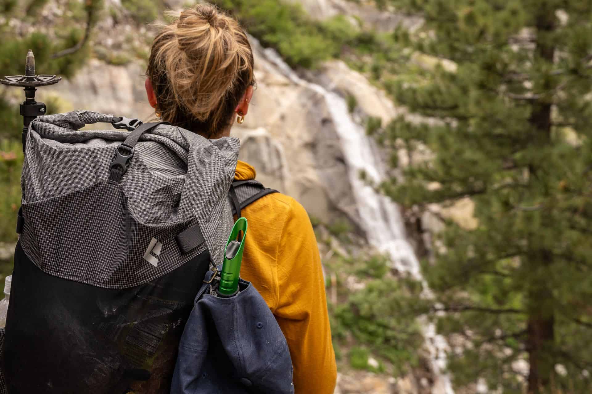 A person with a backpack stands outdoors facing a blurred waterfall and forest. The hiker, perfect for any backpackers Fourth of July sales guide, has blonde hair tied up, wears a yellow jacket, and carries trekking poles and a green water bottle.