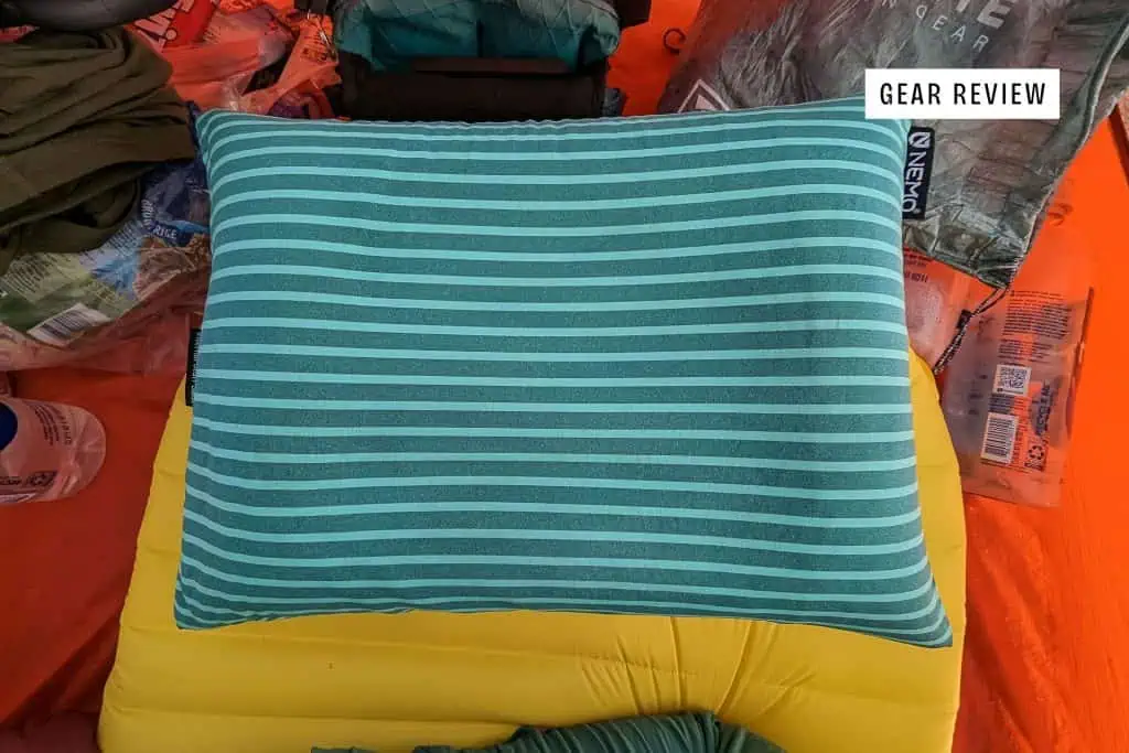 A NEMO Fillo Elite Ultralight Backpacking Pillow with teal and light blue horizontal stripes sits on a yellow inflatable sleeping pad, surrounded by camping gear inside a tent. A label in the upper right reads Gear Review.