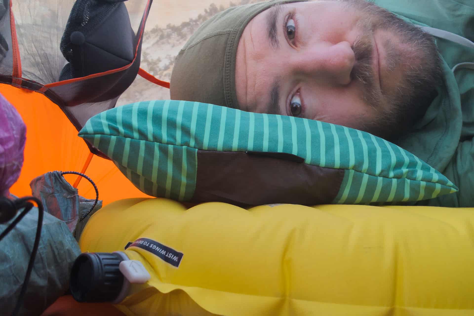 A man lies inside a tent, resting his head on a striped pillow and a yellow sleeping pad, wearing a green hood. Camping gear, including Magma Induction Non-Stick Cookware, is visible around him against the tent’s orange wall.