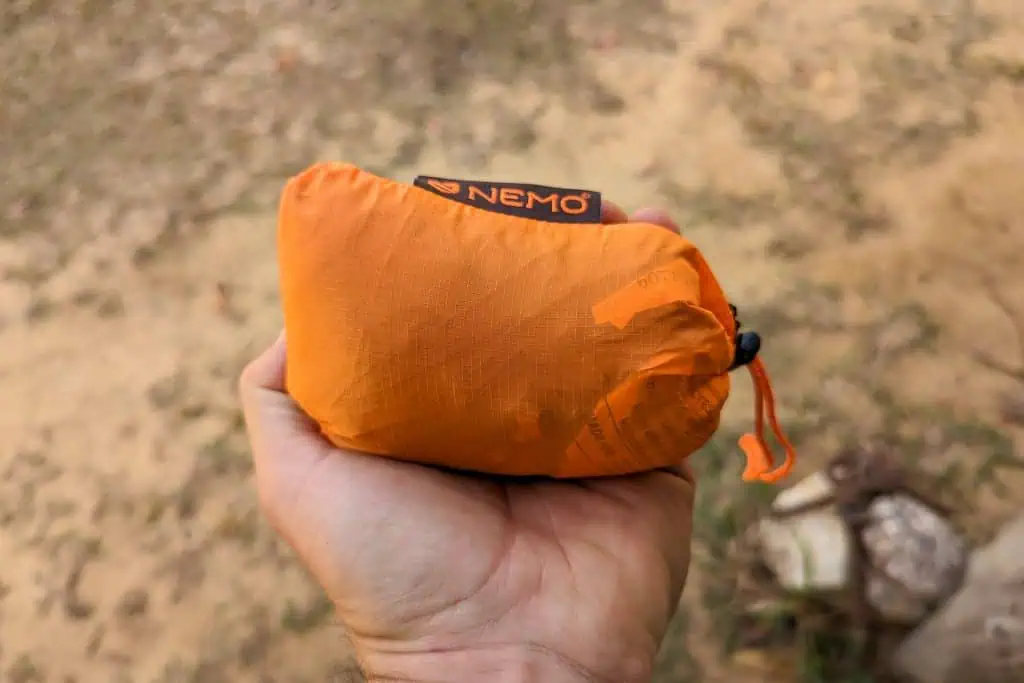 A hand holds a small, orange, compact NEMO Fillo Elite Ultralight Backpacking Pillow pouch with the NEMO logo visible on top. The lightweight pouch looks tightly packed, perfect for outdoor adventures. Dirt and grass are seen in the background.