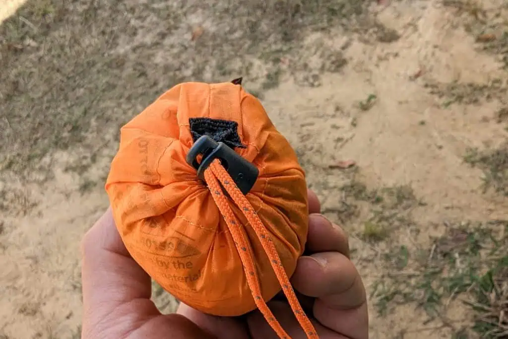 A hand holds a small orange NEMO Fillo Elite Ultralight Backpacking Pillow bag with a black toggle, outdoors on sandy ground with scattered grass.