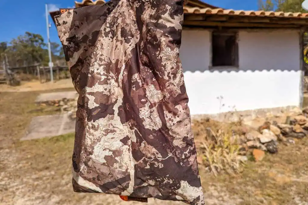 A close-up of a brown and beige camouflage-patterned fabric, possibly part of a NEMO Fillo Elite Ultralight Backpacking Pillow, held outdoors with a rustic white building and dry grass softly blurred in the background.